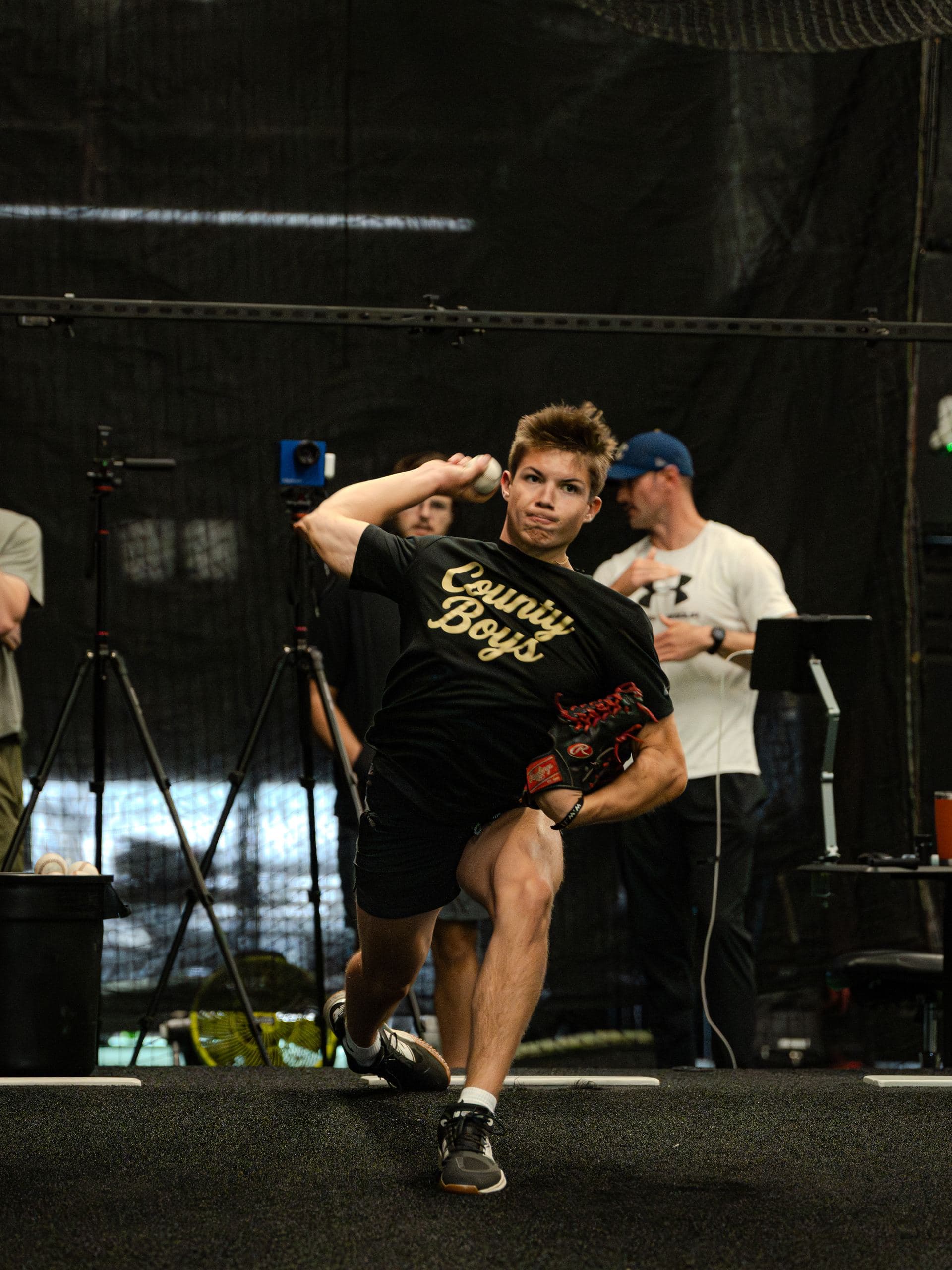 A young pitcher throwing in a PPL tunnel while coaches watch from behind
